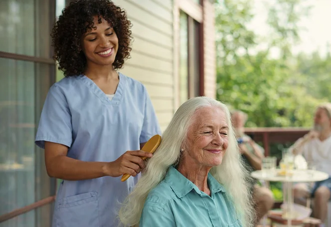 A caregiver brushing an elderly woman's long white hair on an outdoor porch