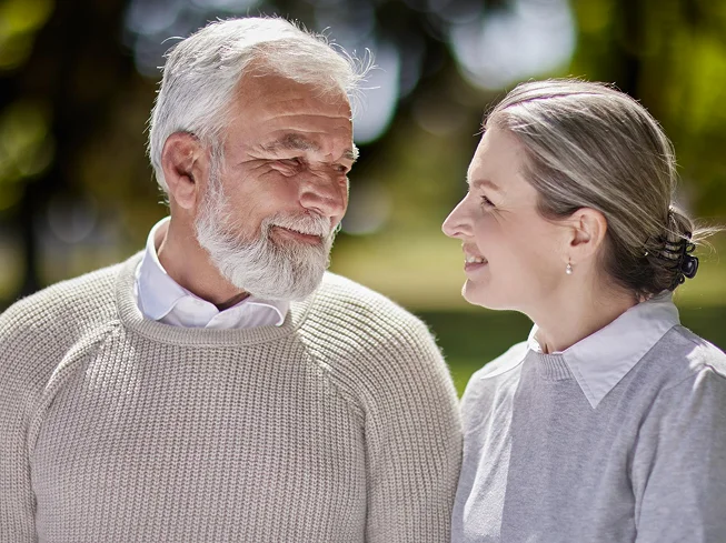 An elderly couple smiling warmly at each other outdoors in a park