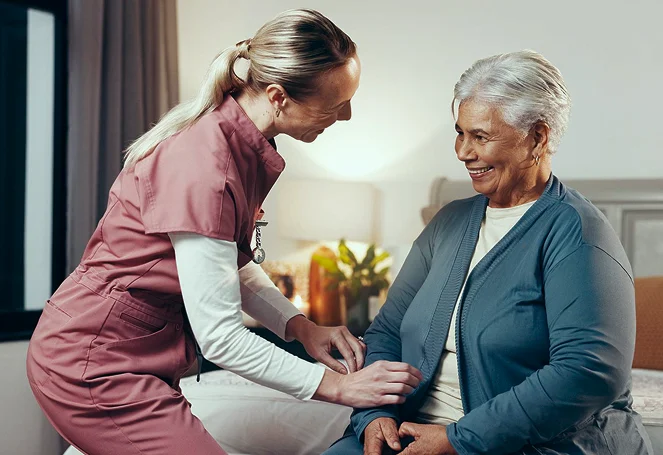 A caregiver attentively assisting a smiling elderly woman seated on a bed