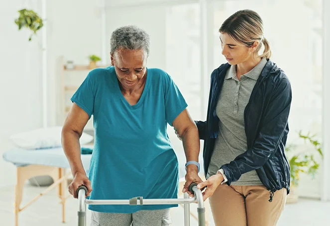 A caregiver supporting an elderly woman walking with a walker indoors