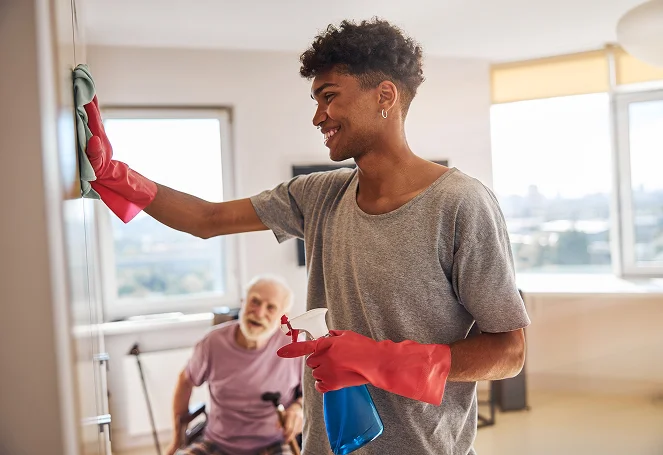 A home aide cleaning a wall with spray bottle while an elderly man sits nearby