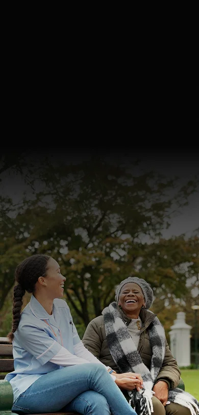 A caregiver and elderly woman laughing together on a park bench in autumn