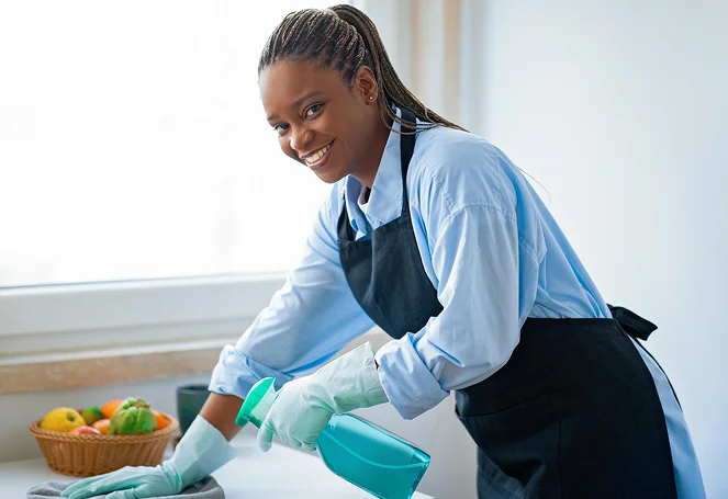 A smiling home aide cleaning a kitchen counter with a spray bottle