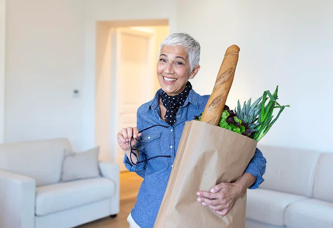 A cheerful senior woman carrying a paper bag of fresh groceries at home