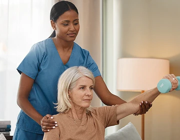 Caregiver in blue scrubs assisting an elderly woman with a dumbbell arm exercise during a home rehabilitation session
