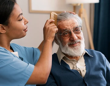 Female caregiver in blue scrubs combing the hair of a smiling elderly man during a home care visit