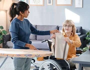 Caregiver helping an elderly woman in a wheelchair with folding laundry in a home living room