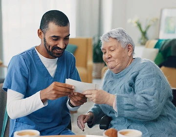 Male caregiver in blue scrubs reviewing a document with an elderly woman at a table during a home care visit