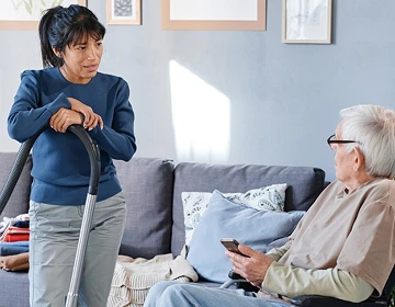 Home caregiver pausing while vacuuming to talk with an elderly man sitting on a sofa in a living room