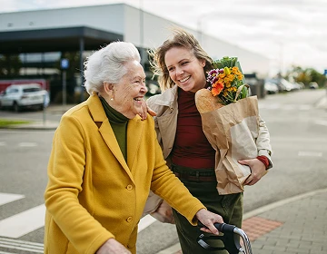 Caregiver walking and laughing with an elderly woman using a walker after grocery shopping outdoors