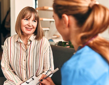 Smiling elderly woman talking with a nurse taking notes on a clipboard during a home care assessment