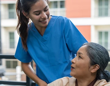 Smiling caregiver in blue scrubs attending to an elderly woman in a wheelchair on an outdoor balcony