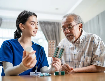 Female caregiver in blue scrubs explaining medication to an elderly man at a table during a home care visit