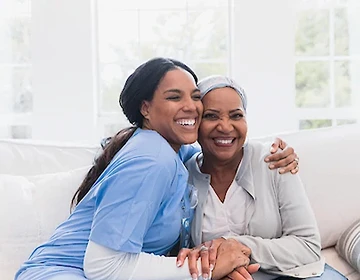 Smiling caregiver in blue scrubs hugging a happy elderly woman on a sofa in a bright living room