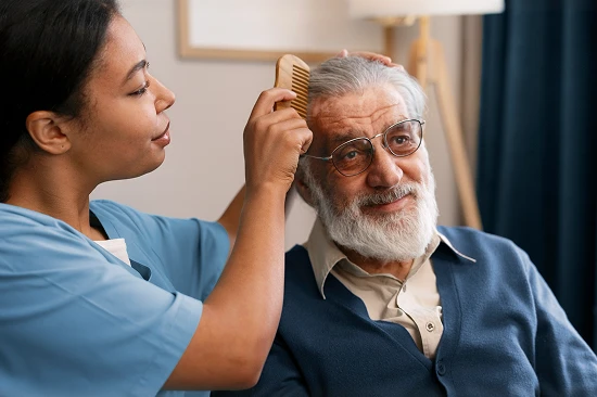 Female caregiver in blue scrubs combing the hair of a smiling elderly man during a home care visit