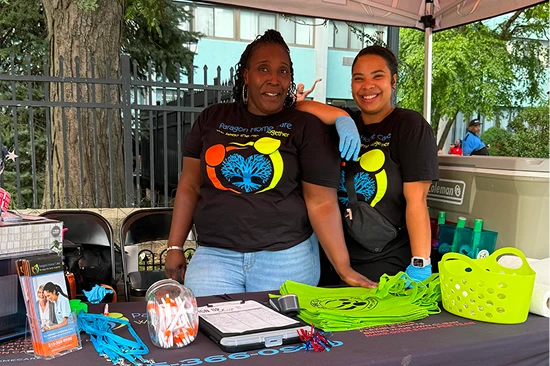 Two Paragon Home Care staff members smiling at their booth table with branded giveaways at an outdoor community event