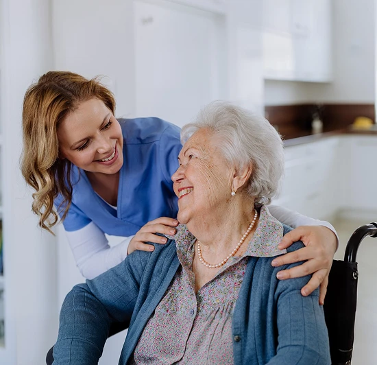 Smiling caregiver in blue scrubs embracing a happy elderly woman in a wheelchair at home