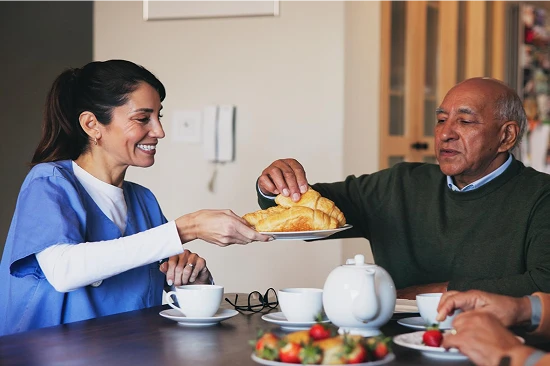 Smiling caregiver in blue scrubs serving a croissant to an elderly man at a breakfast table during a home care visit