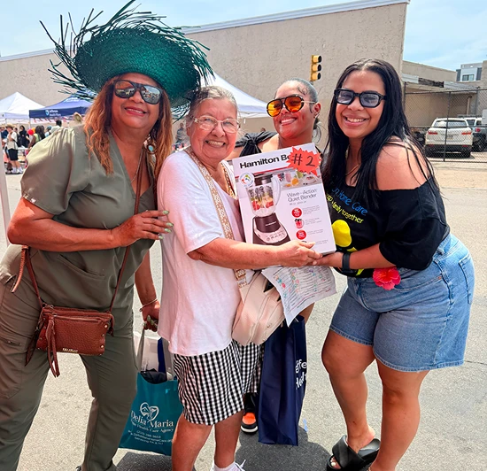 Group of four women smiling outdoors at a community event as an elderly woman holds a Hamilton Beach blender prize