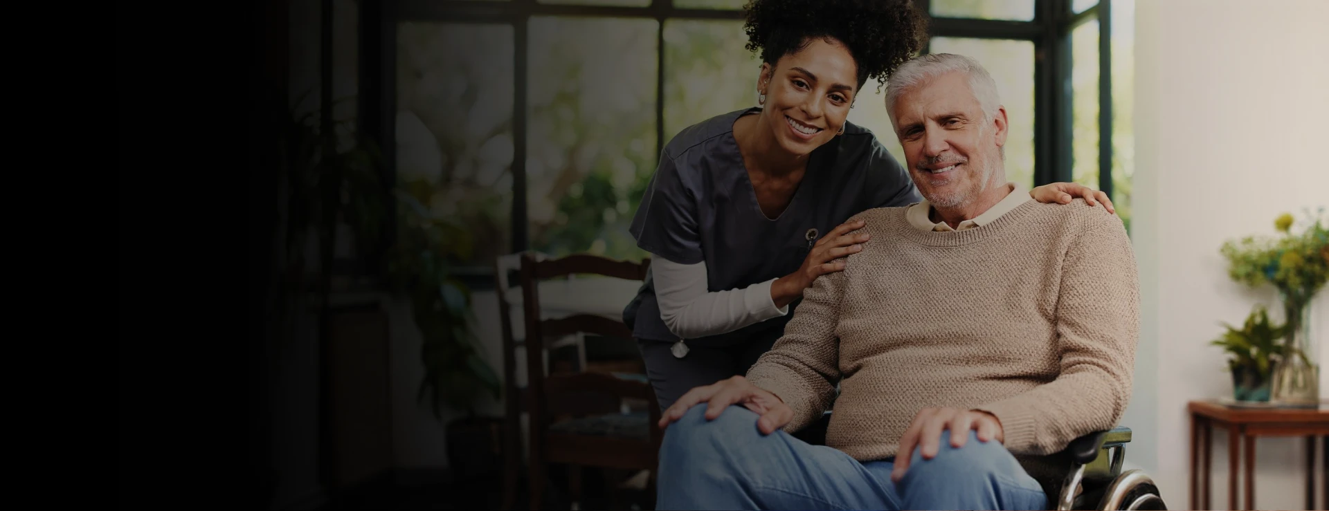 Smiling caregiver in dark scrubs posing with a cheerful elderly man in a wheelchair in a bright home setting