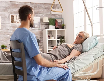 Male caregiver in blue scrubs sitting beside an elderly woman resting in an adjustable bed at home