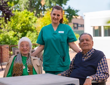 Caregiver in green scrubs standing between a smiling elderly woman and man seated at an outdoor table