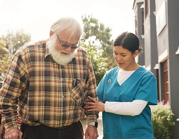 Caregiver in blue scrubs steadying an elderly man in a plaid shirt as they walk outdoors
