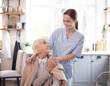 Caregiver in light blue scrubs resting a hand on a smiling elderly woman's shoulder in a bright home kitchen