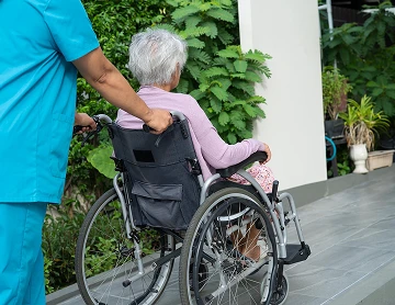 Caregiver in blue scrubs pushing an elderly woman in a wheelchair along a walkway beside green plants