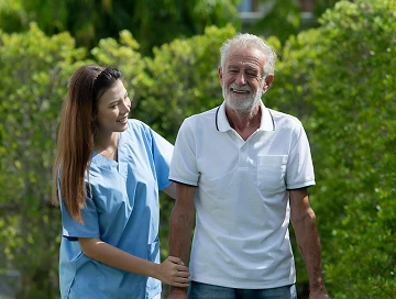 Smiling caregiver in blue scrubs supporting an elderly man's arm as they walk together outdoors