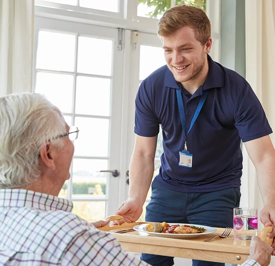 Smiling male caregiver serving a meal tray to an elderly man seated by a bright window