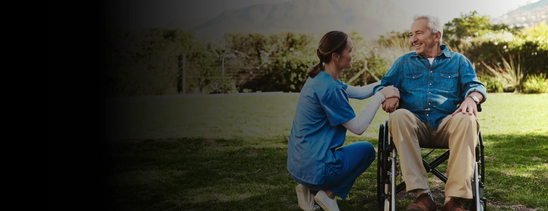 Caregiver in blue scrubs kneeling beside a smiling senior man in a wheelchair, holding his hand in a sunny garden